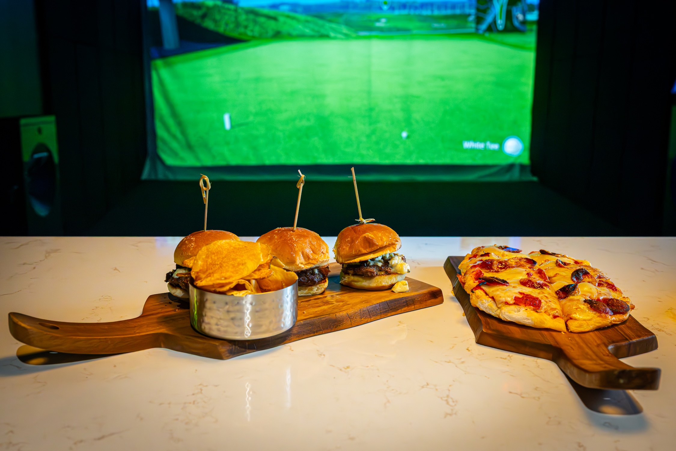 bar top with flatbread pizza and burger sliders in front of a golf simulator bay at ParT Barn