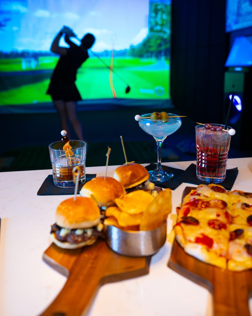 a person mid-swing in a golf simulator and a bar top with food and drinks behind them at ParT Barn